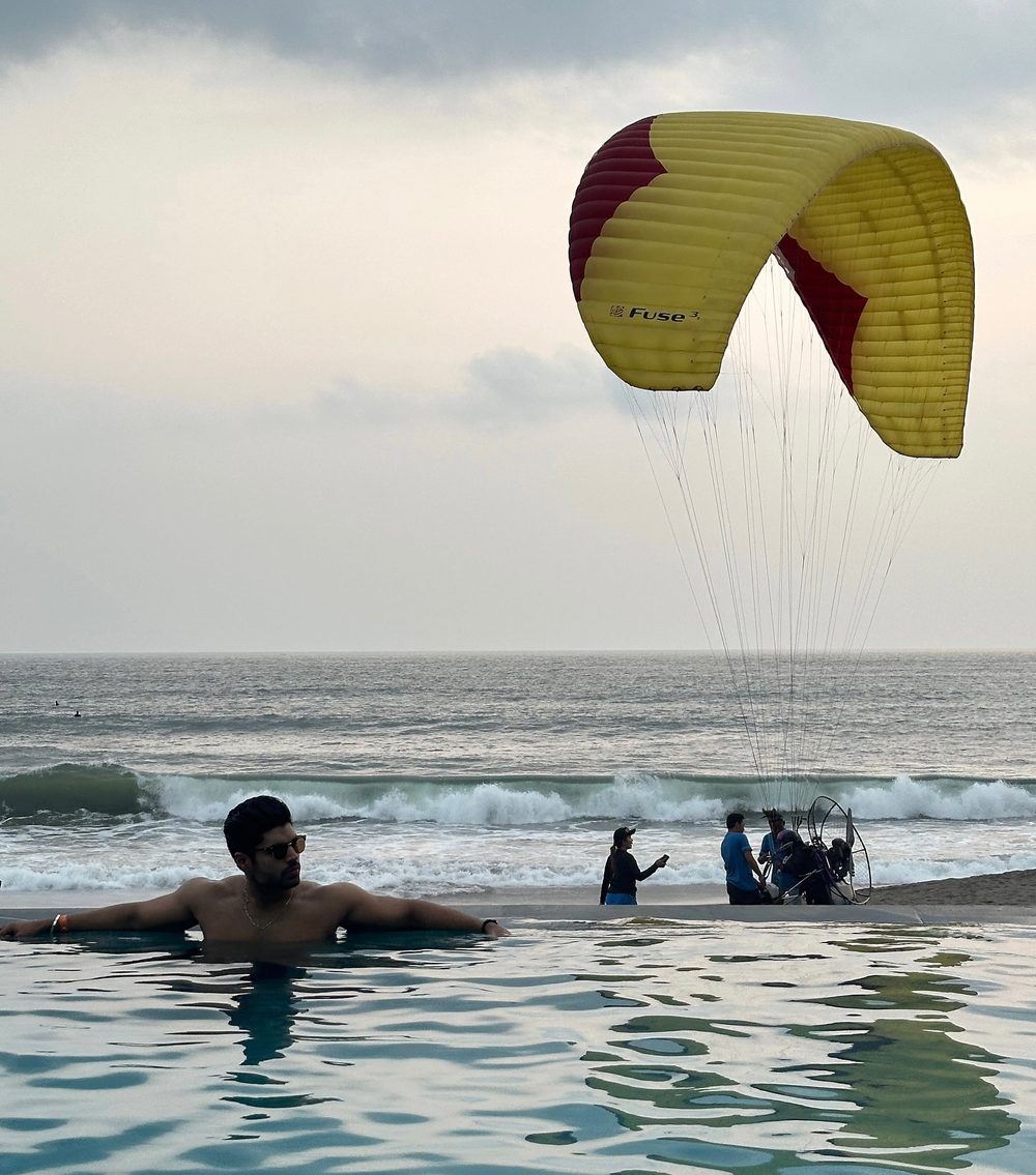 Man in an infinity pool overlooking a beach with a paraglider landing